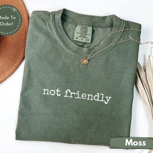 May include: A moss green t-shirt with the phrase "not friendly" printed in white. The shirt has a crew neck and short sleeves. A gold necklace with a pendant is draped across the shirt.
