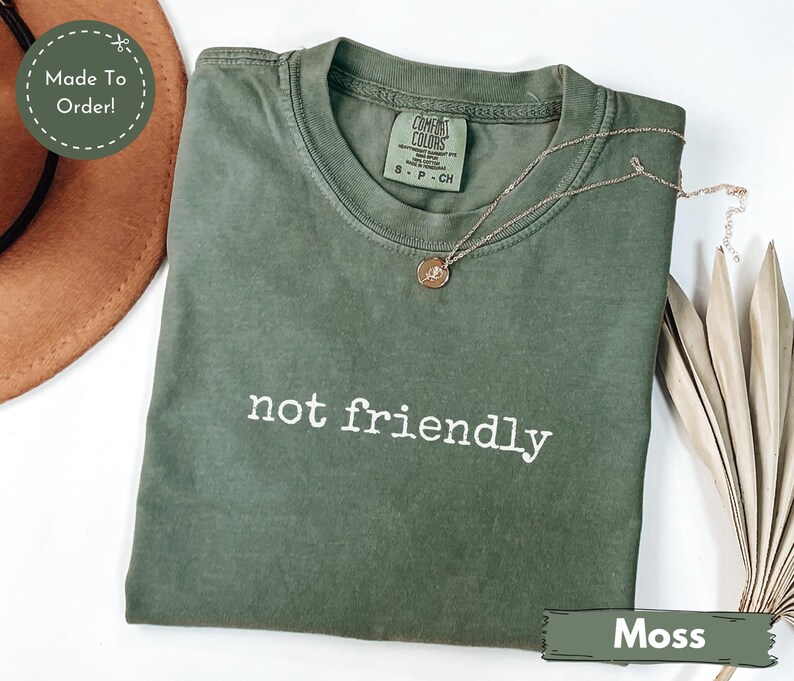 May include: A moss green t-shirt with the phrase "not friendly" printed in white. The shirt has a crew neck and short sleeves. A gold necklace with a pendant is draped across the shirt.