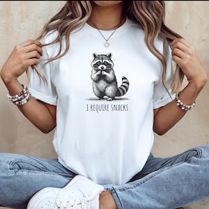Könnte beinhalten: Weißes T-Shirt mit einer schwarz-weißen Illustration eines Waschbären. Der Waschbär sitzt und hält seine Pfoten an seinem Mund. Unter der Illustration steht der Text "I REQUIRE SNACKS".