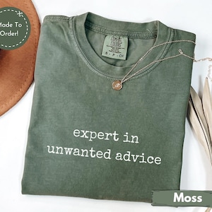 May include: A moss green t-shirt with the text "expert in unwanted advice" in white. The shirt has a crew neck and short sleeves. A gold necklace with a pendant is draped on the shirt.