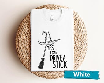 Può includere: T-shirt bianca con grafica nera di cappello da strega e scopa, con la scritta "YES I CAN DRIVE A STICK". La maglietta è esposta su una superficie rotonda e intrecciata. Un banner blu in basso recita "White".