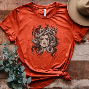 May include: Orange t-shirt featuring a Medusa graphic. The illustration depicts a woman with snakes for hair, with a distressed, vintage style. The shirt is made of a soft material and has a classic crew neck.