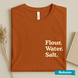 May include: A rust-colored t-shirt with the words "Flour. Water. Salt." in a white, sans-serif font. The shirt has a crew neck and a small white tag on the collar. The word "Autumn" is written on a blue banner at the bottom.