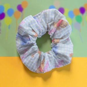 May include: A light blue scrunchie with a plaid pattern and cartoon bear and elephant illustrations. The hair accessory is made of soft fabric and is designed to hold hair in a ponytail or bun. The scrunchie is a cute and practical hair accessory.