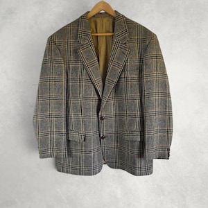 Op de afbeelding: Een vintage blazer met een klassiek ruitpatroon in bruin, zwart en beige. De jas heeft een klassiek ontwerp met een revers, twee voorzakken en een sluiting met twee knopen. Het hangt aan een houten kleerhanger tegen een neutrale achtergrond.