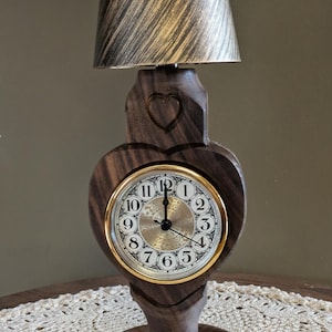 May include: A wooden table lamp with a heart-shaped clock face. The clock has a gold-colored bezel and black numerals. The lamp shade is a light brown color with a textured pattern. The lamp is on a white lace tablecloth.