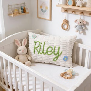 May include: A white crib with a plush bunny and a pillow that says "Riley" in green yarn. The pillow has a textured surface and is decorated with small flower accents. A wooden rattle is on the mattress.