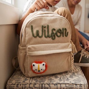 Puede incluir: Una mochila de pana beige con el nombre Wilson bordado en hilo verde oscuro en la parte superior. El bolsillo delantero muestra un pequeño parche redondo de felpa con forma de cara de león, con una melena naranja brillante y detalles.