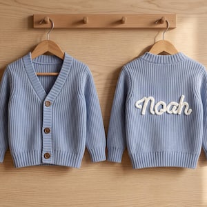 May include: Two light blue knit cardigans with long sleeves and ribbed textures. The left cardigan features a V-neckline with three brown buttons. The right cardigan displays the name Noah in white cursive lettering embroidered on the back.