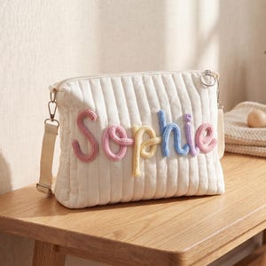 Puede incluir: Un bolso cruzado acolchado blanco con el nombre "Sophie" bordado en hilo colorido. El bolso tiene una correa beige y una cremallera plateada. Se exhibe en un banco de madera.