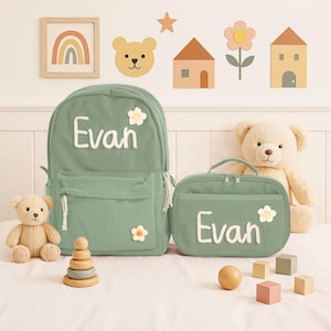 May include: A sage green backpack and matching lunch bag with the name "Evan" in white script. Both items have a daisy accent. A teddy bear and wooden toys are also in the image. The backpack has a front pocket and a white cord zipper.
