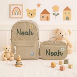 May include: A beige corduroy backpack and lunch bag set with the name "Noah" embroidered in green. The backpack has a small flower applique. A teddy bear and wooden toys are also in the image. Decorative wall art includes a rainbow, bear, houses, and a star.
