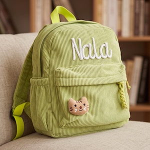 May include: A light green corduroy backpack featuring the name Nala embroidered in white script on the front pocket. The bag includes a bright lime green top handle and shoulder straps, along with a small, plush tan cat face patch attached.