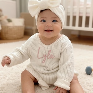 Könnte beinhalten: Ein Baby in einem cremefarbenen Strick-Outfit, bestehend aus einem langärmeligen Oberteil und Shorts, mit dem Namen "Lyla" in Rosa gestickt. Das Outfit wird durch ein passendes Schleifen-Stirnband ergänzt. Das Baby sitzt auf einem cremefarbenen Teppich.