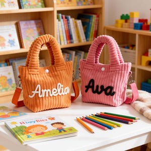 May include: Two corduroy tote bags, one orange and one pink, sit on a white table. The orange bag has the name "Amelia" embroidered on it, and the pink bag has "Ava." Books and colored pencils are also on the table. Shelves filled with books are in the background.