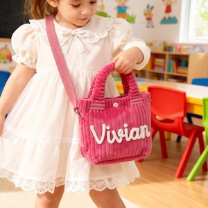 May include: A young girl wearing a white dress with a pink shoulder bag. The bag is corduroy with the name "Vivian" in white script. The bag has a button closure and a matching pink strap.
