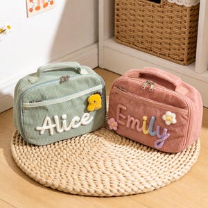 May include: Two corduroy lunch bags, one mint green and one dusty rose, sit on a woven mat. The mint bag has "Alice" in white letters and a yellow duck. The rose bag has "Emily" and flower accents.