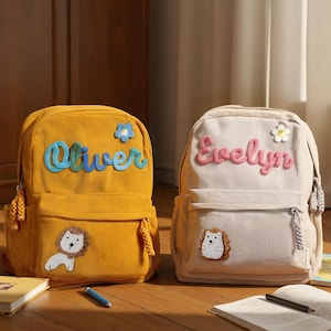 May include: Two corduroy backpacks, one mustard yellow and one beige. The yellow backpack has the name "Oliver" in blue script and a lion patch. The beige backpack has the name "Evelyn" in pink script and a hedgehog patch.