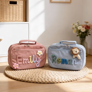 May include: Two personalized corduroy bags, one pink and one blue, sit on a woven mat. The pink bag has the name "Emily" in colorful letters and a flower. The blue bag features the name "Roman" and a lion applique.