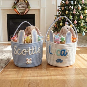 May include: Two Easter baskets, one light blue and one white, with bunny ear accents and handles. The blue basket says "Scottie" and has a bunny applique. The white basket says "Lucas". Both are filled with Easter eggs and a stuffed bunny.