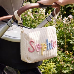 May include: A cream-colored quilted stroller bag featuring the name Sophie in raised, multi-colored yarn lettering. The bag is attached to the brown leather-look handle of a stroller with two cream fabric straps and gold-toned metal clips.