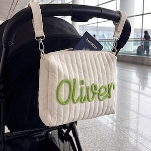 Peut inclure: Sac de poussette matelassé crème avec le nom "Oliver" brodé en vert. Le sac est suspendu à une poignée de poussette noire et contient un passeport. Le sac est rectangulaire avec un motif matelassé vertical.