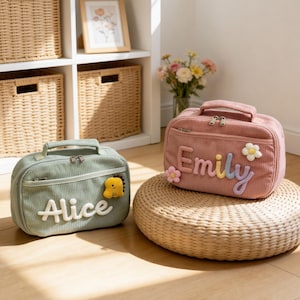 May include: Two corduroy lunch bags, one sage green and one dusty rose, with embroidered names "Alice" and "Emily". Each bag has a top handle and a zipper closure. The "Emily" bag rests on a woven stool.