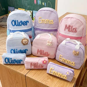 May include: A collection of striped backpacks, lunch bags, and pencil cases in blue, purple, and pink. Each item is personalized with embroidered names including Oliver, Emma, and Mila, and features small plush animal or flower accents.