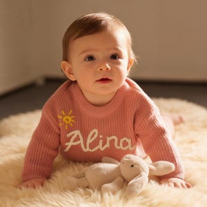 Puede incluir: Un bebé con un suéter rosa con el nombre "Alina" bordado en blanco y un diseño de sol amarillo. Un conejito de peluche crema descansa sobre una alfombra esponjosa. El bebé mira a la cámara.
