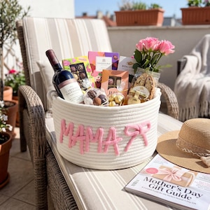May include: A white rope basket with the word "MAMA" in pink yarn, filled with a bottle of wine, chocolates, cookies, and pink roses. A straw hat and a "Mother's Day Gift Guide" magazine are nearby.