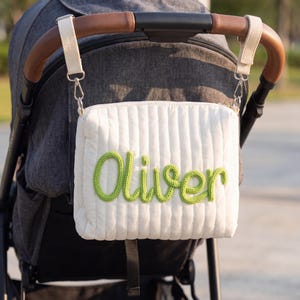 May include: A white, quilted stroller organizer bag with the name "Oliver" embroidered in green. The bag is attached to a gray stroller with a brown leather handle. The bag has a rectangular shape and a vertical ribbed pattern.