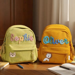 May include: Two corduroy backpacks, one light green and one yellow. The light green backpack has the name "Sophie" embroidered in colorful thread, and a cat patch. The yellow backpack has the name "Oliver" embroidered in blue thread, and a lion patch.