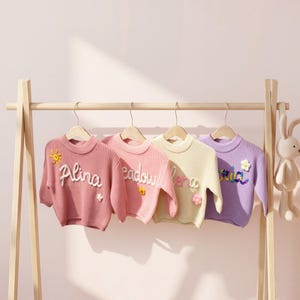 Op de afbeelding: Vier pastelkleurige kindertruien hangen aan een houten kledingrek. Elke trui heeft een andere kleur: roze, lichtroze, geel en paars. De truien hebben geborduurde namen en bloemdetails. Een knuffelkonijn hangt aan de zijkant.