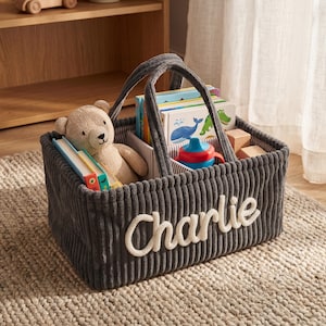 May include: A grey corduroy storage basket with the name "Charlie" embroidered in white. The basket contains books, a teddy bear, and a sippy cup. The basket has a handle for easy carrying.
