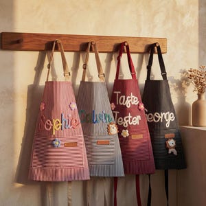 Może przedstawiać: Cztery paski w paski wiszące na drewnianym wieszaku. Każdy fartuch ma inny schemat kolorów i zawiera imię lub frazę w ozdobnych literach. Fartuchy mają kieszenie i kwiatowe lub zwierzęce zdobienia.