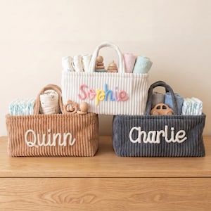 May include: Three corduroy baby baskets in white, brown, and navy blue, each with a handle and a name embroidered on the front: "Sophie", "Quinn", and "Charlie". The baskets are filled with baby essentials and toys.