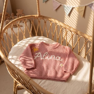 May include: A pink knitted baby sweater with the name "Alina" embroidered in white script. The sweater features a small yellow sun and flower embellishments. The sweater is displayed on a white cushion inside a wicker cradle.