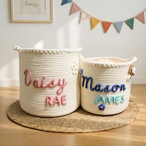 May include: Two off-white rope storage baskets. One basket has the name "Daisy Rae" in pink lettering, and the other has "Mason James" in blue and teal lettering. Both baskets have decorative flowers.
