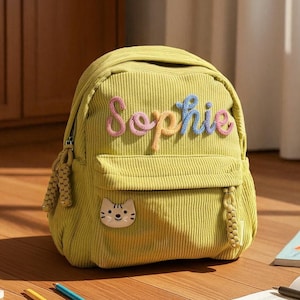 May include: A yellow corduroy backpack with the name "Sophie" embroidered in colorful thread. The backpack has a front pocket with a cat face patch and a top handle. The bag is sitting on a wooden surface.