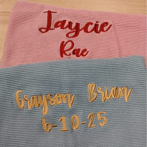 Pode incluir: Duas mantas de malha, uma rosa e outra azul. A manta rosa tem os nomes "Jaycie Rae" bordados em vermelho. A manta azul tem "Grayson Brian" e a data "6-10-25" bordados em dourado.