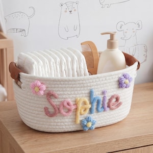 May include: An oval white cotton rope basket featuring brown leather handles and the name Sophie spelled out in colorful yarn letters. The basket is decorated with small pink, purple, and blue yarn flowers and contains white diapers and a comb.