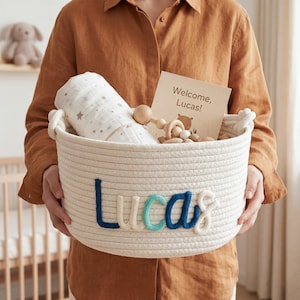 May include: A white woven rope basket featuring the name Lucas in blue and teal yarn lettering. Inside the basket is a white swaddle blanket with a star pattern, a wooden bead rattle, and a card with the text Welcome, Lucas! and a bear icon.