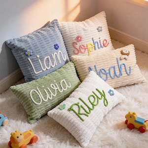 Peut inclure: Cinq coussins décoratifs en velours côtelé de différentes couleurs, chacun brodé d'un nom. Les coussins sont bleu clair, crème, vert et blanc, avec des noms comme "Liam", "Sophie", "Olivia", "Noah" et "Riley". Chaque coussin a des accents floraux.