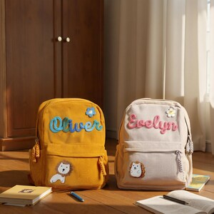 May include: Two corduroy backpacks, one mustard yellow and one beige. The yellow backpack has the name "Oliver" in blue script and a lion patch. The beige backpack has the name "Evelyn" in pink script and a hedgehog patch.