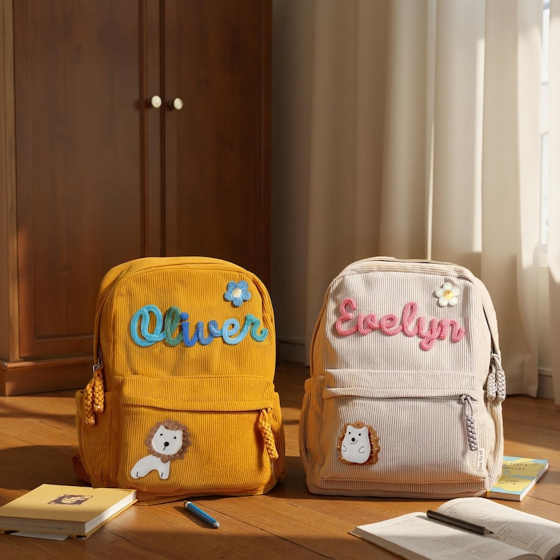 May include: Two corduroy backpacks, one mustard yellow and one beige. The yellow backpack has the name "Oliver" in blue script and a lion patch. The beige backpack has the name "Evelyn" in pink script and a hedgehog patch.