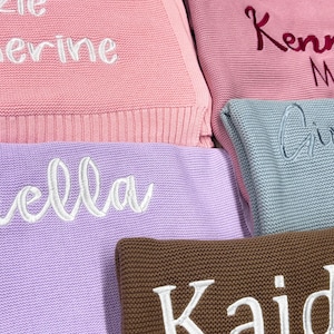 Pode incluir: Close-up de v&aacute;rias mantas de malha em v&aacute;rias cores pastel, incluindo rosa, lavanda e azul. Cada manta apresenta um nome bordado em uma cor contrastante. Os nomes est&atilde;o em uma fonte cursiva, adicionando um toque personalizado.