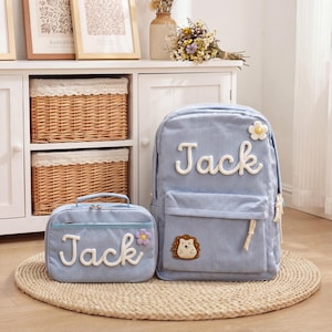 May include: Light blue corduroy backpack and lunch bag set. The backpack and lunch bag have the name "Jack" in white script. The backpack features a flower and hedgehog patch. The lunch bag has a flower patch. The set is on a woven mat.