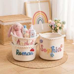 May include: Two off-white woven rope storage baskets featuring braided rims and colourful yarn lettering. One basket displays the name Amelia with small flowers, the other shows Roman with a red car, and a third shows Sophia with blue flowers.