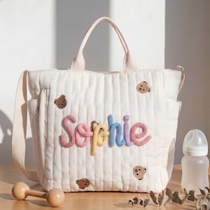 May include: A cream-colored quilted tote bag featuring the name Sophie in raised, multi-colored yarn lettering. The bag is decorated with small embroidered brown teddy bear faces and includes two top handles and a side shoulder strap.