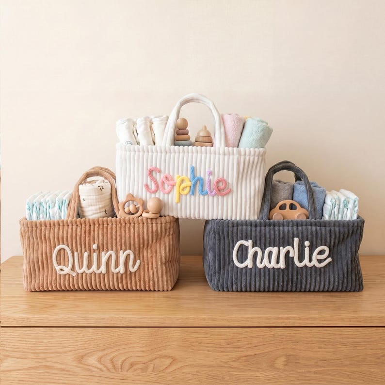 May include: Three corduroy baby baskets in white, brown, and navy blue, each with a handle and a name embroidered on the front: "Sophie", "Quinn", and "Charlie". The baskets are filled with baby essentials and toys.